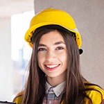 A woman with long brown hair, wearing a yellow construction helmet and a plaid shirt, smiles while standing indoors near a gray wall.